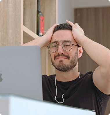 Frustrovaný muž s okuliarmi si drží hlavu oboma rukami pri pohľade na notebook.